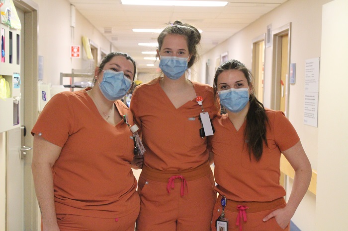 Three women in orange scrubs and wearing masks with arms around each other.