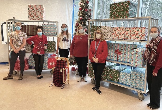 Six women standing in front of two three-shelf units full of wrapped gifts.