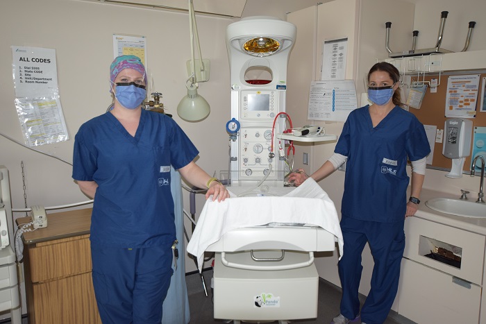 Two women in blue scrubs and masks stand beside a large white piece of machinery that has a baby bed attached.