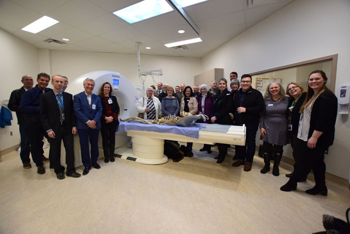 A large group if people gather around a large white round machine that has a bed attached. There is a medical skeleton on the bed.