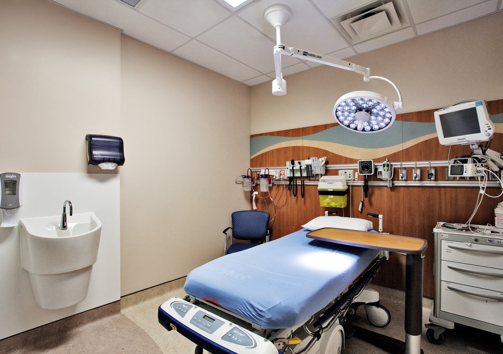 A hospital room with a bed, sink, crash cart and overhead operating light.