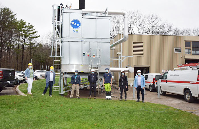 Seven people wearing hardhats stand in front of the new large metal cooling tower outside GBGH