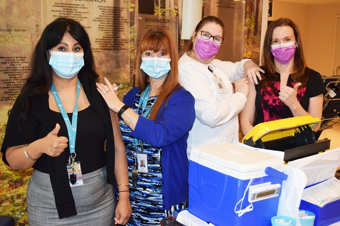 Four women wearing masks standing behind a blue cooler containing flu shot vaccines.