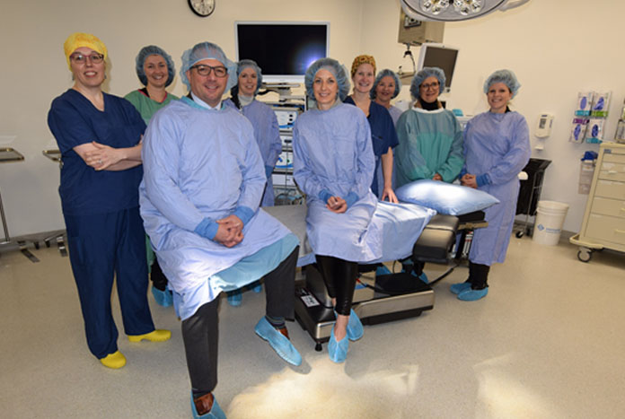 A group of nine people pose gathered around an operating room bed. They are all wearing gowns, scrubs and head coverings.