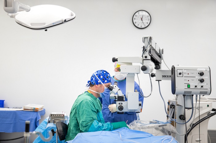 A doctor looking through an eyepiece on a machine as he operates. An assistant is beside him.