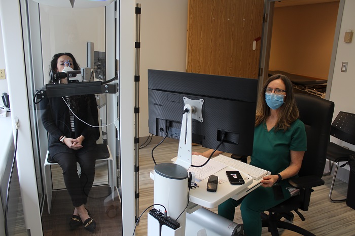 Female medical person running a computer while a patient sits in a glass booth with a breathing apparatus on her face.