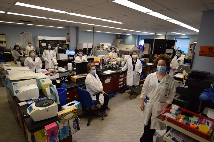 A large room with wooden desks and equipment. Twelve people in masks and lab coats stand a minimum of 6 feet apart.