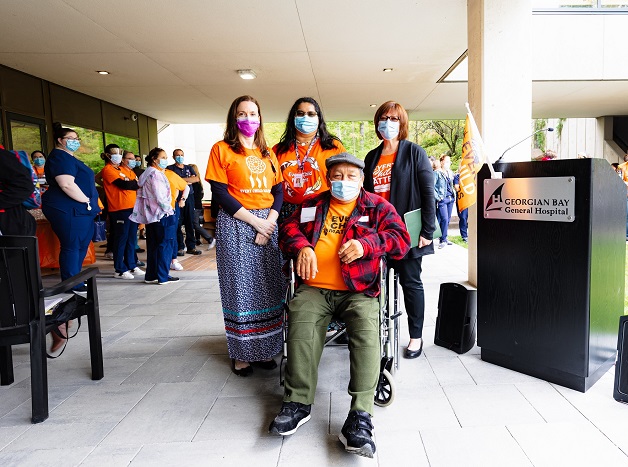 A man in wheel chair with three women standing behind. A large group of people are in the background. Most are wearing orange t-shirts.