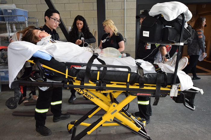 A medical responder wheels in a woman on a yellow stretcher outside of the Emergency entrance.