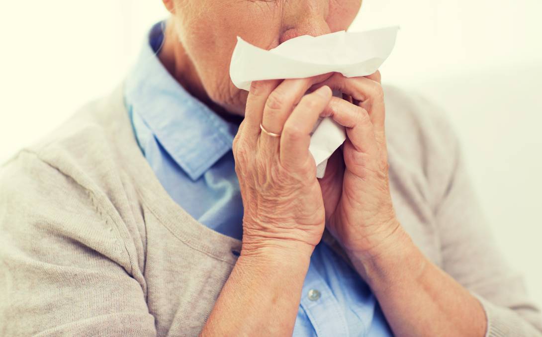 An older person wearing a gold wedding band blows their nose with a tissue.